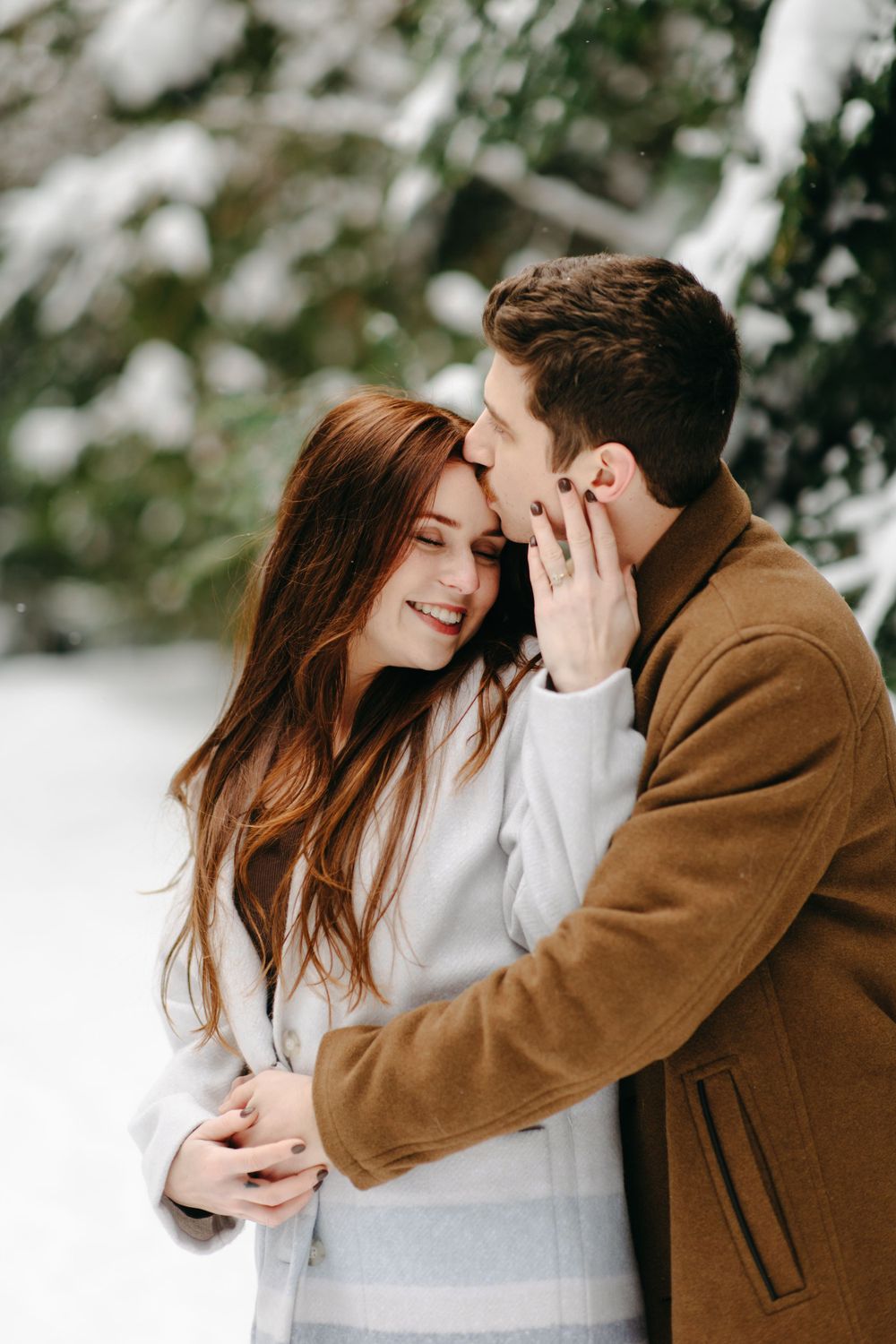 A romantic winter couple embracing outdoors in warm coats against a snowy forest background.