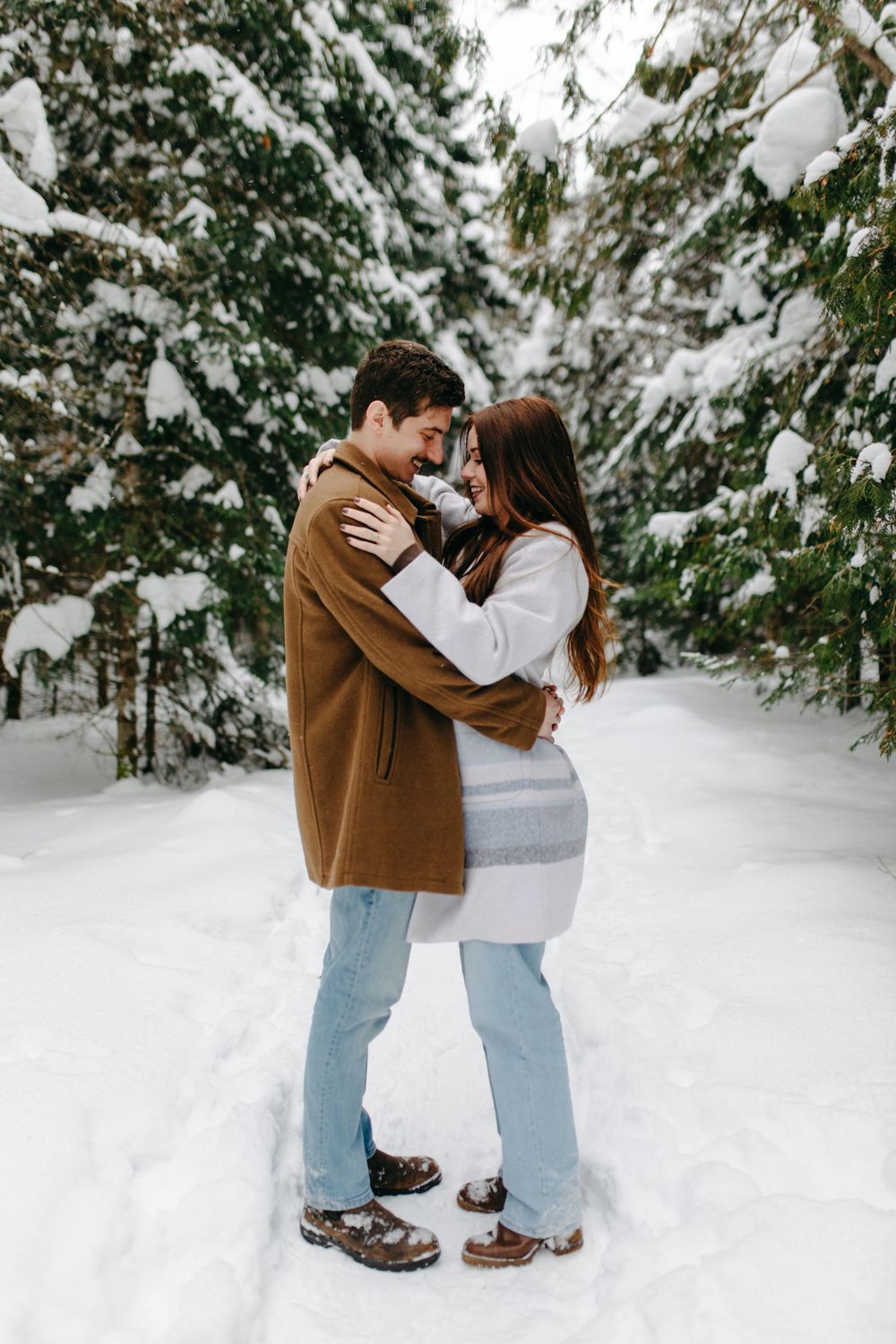 A couple embraces in a snow-covered forest setting wearing winter clothing against evergreen trees and fresh snowfall.