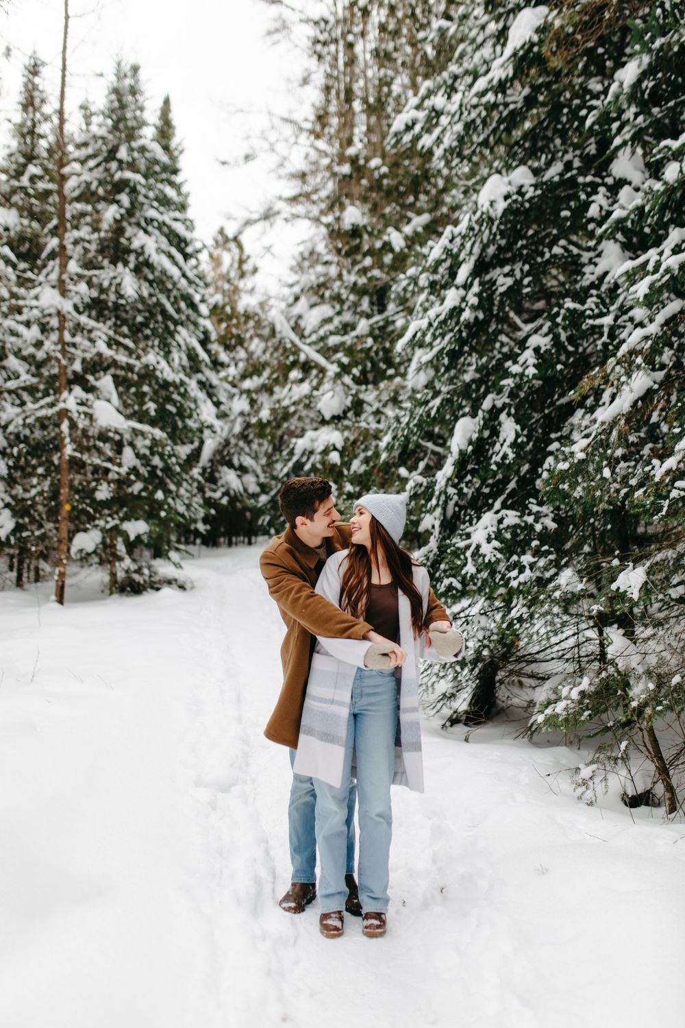 A couple shares an intimate moment on a snow-covered forest path surrounded by evergreen trees dusted with fresh snow.