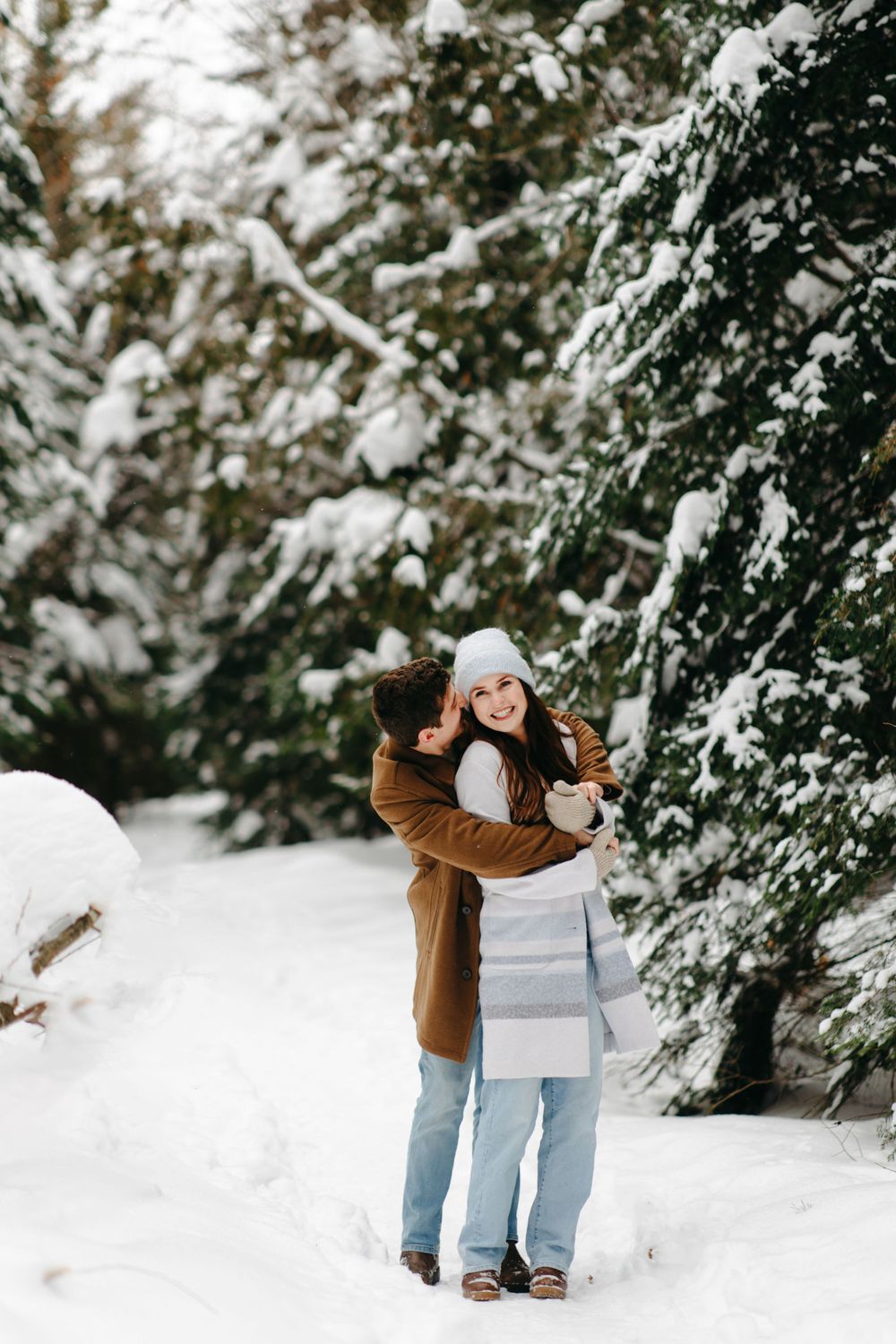 A couple embraces in a snowy winter forest scene with snow-covered evergreen trees in the background.