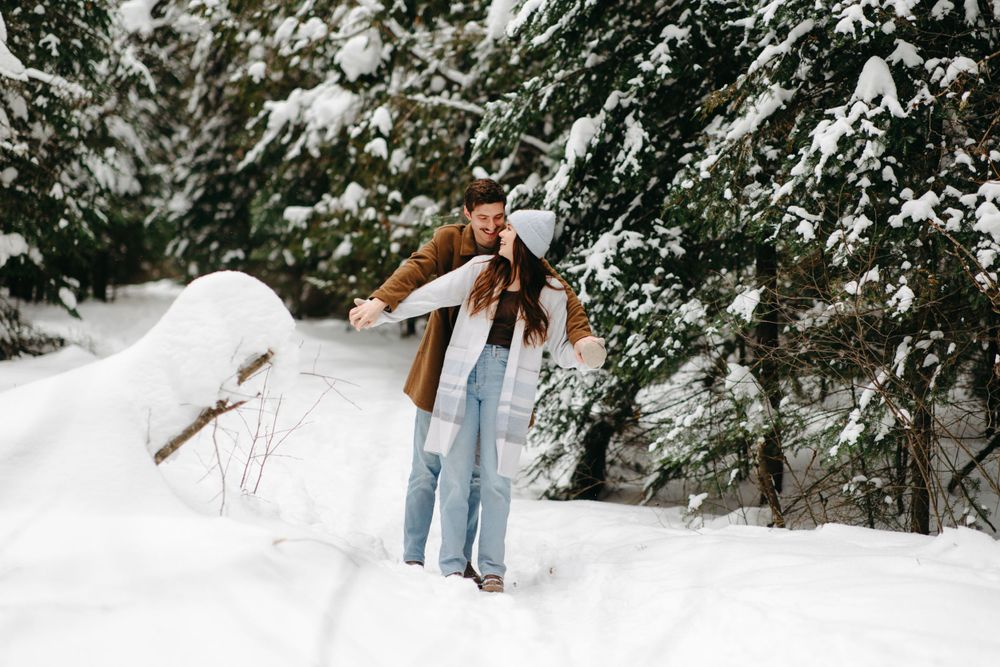 A person in a brown coat and jeans stands in a snowy forest with snow-covered evergreen branches overhead.