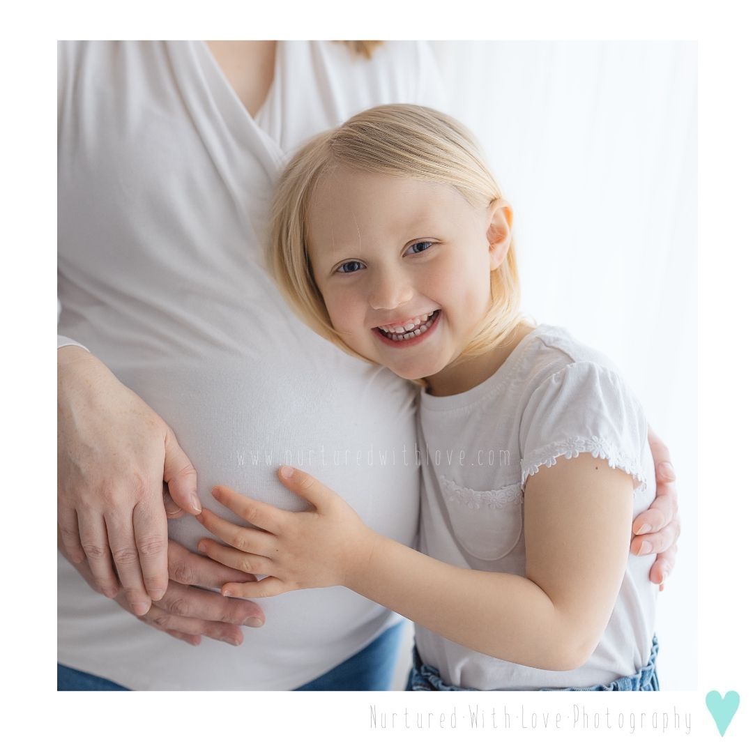 A joyful young child in a white shirt embraces a pregnant woman's belly with excitement.