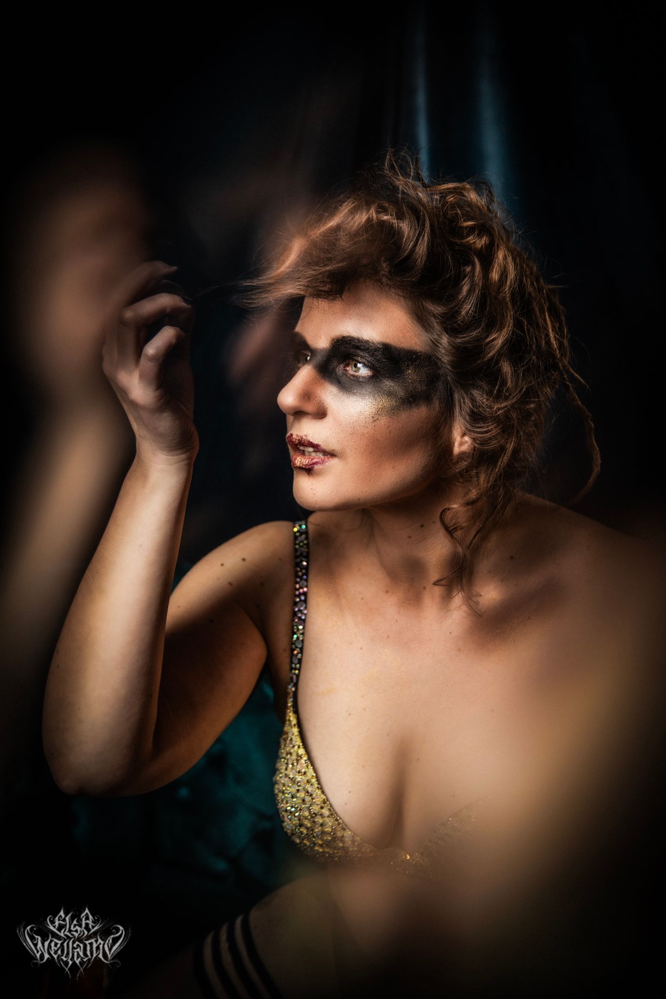 Glamorous portrait with dramatic lighting features bold eye makeup, tousled hair, and a gold sequined top in a moody setting.