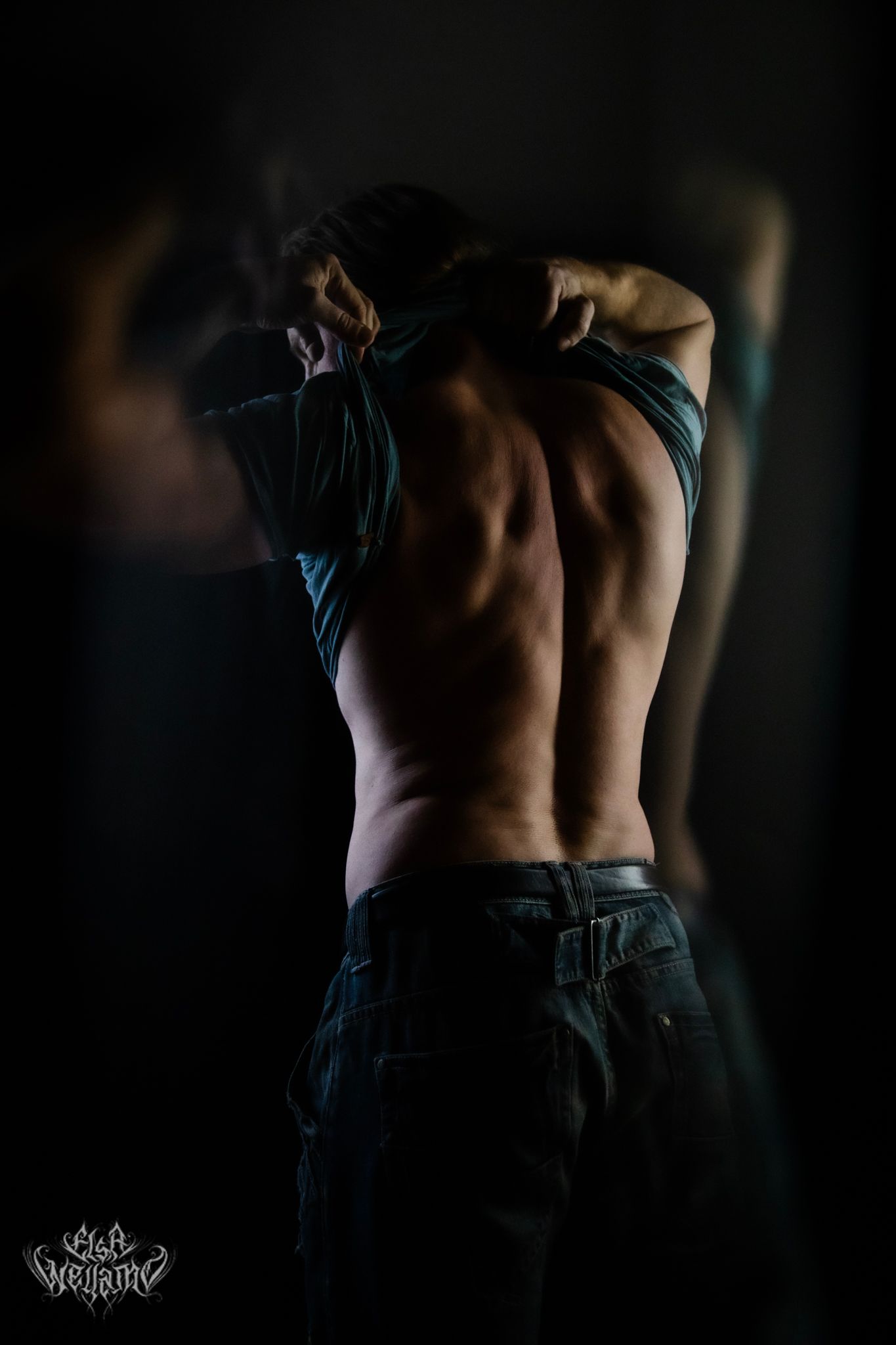 Dramatic low-key artistic portrait of shirtless person removing clothing in dark moody lighting.