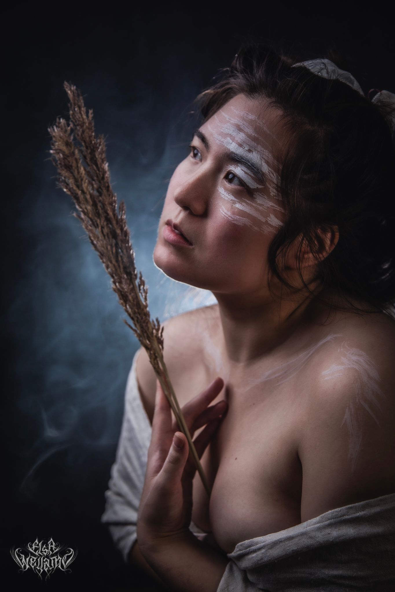 Ethereal artistic portrait with white face paint and dried pampas grass against dramatic dark background.