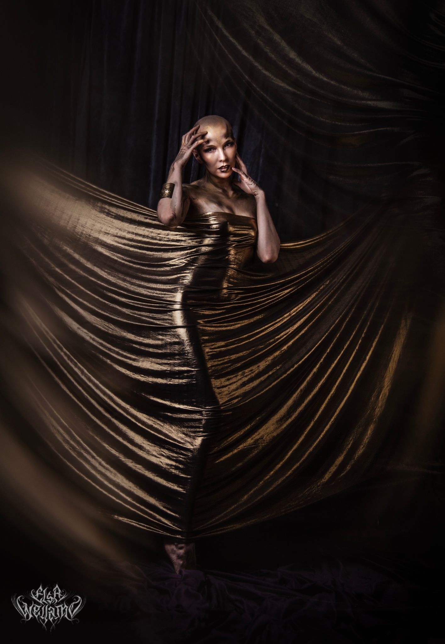 Artistic portrait against dark background with dramatic gold fabric creating flowing lines and shadow patterns.