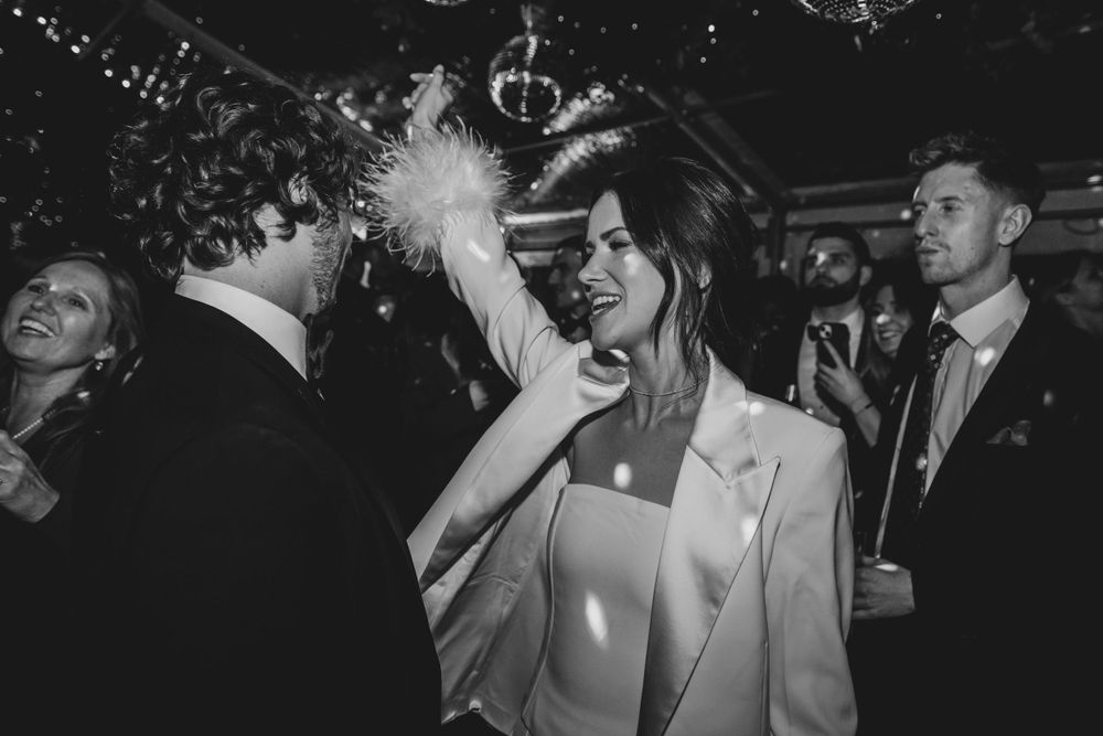 A black and white photo captures a bride in a white suit dancing joyfully at her wedding reception.