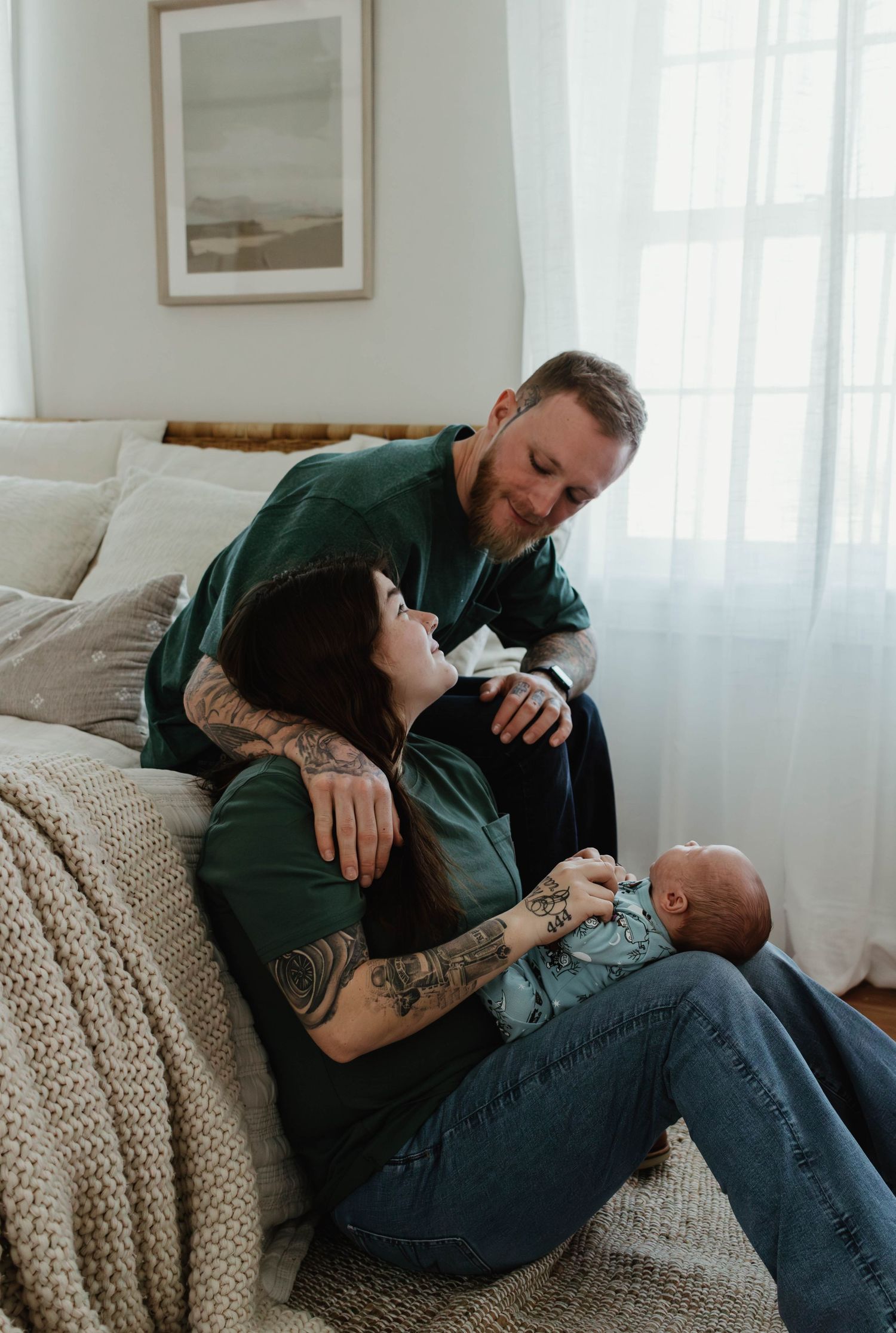 A couple in matching green shirts cuddles on a couch while bonding with their newborn in a bright living room.