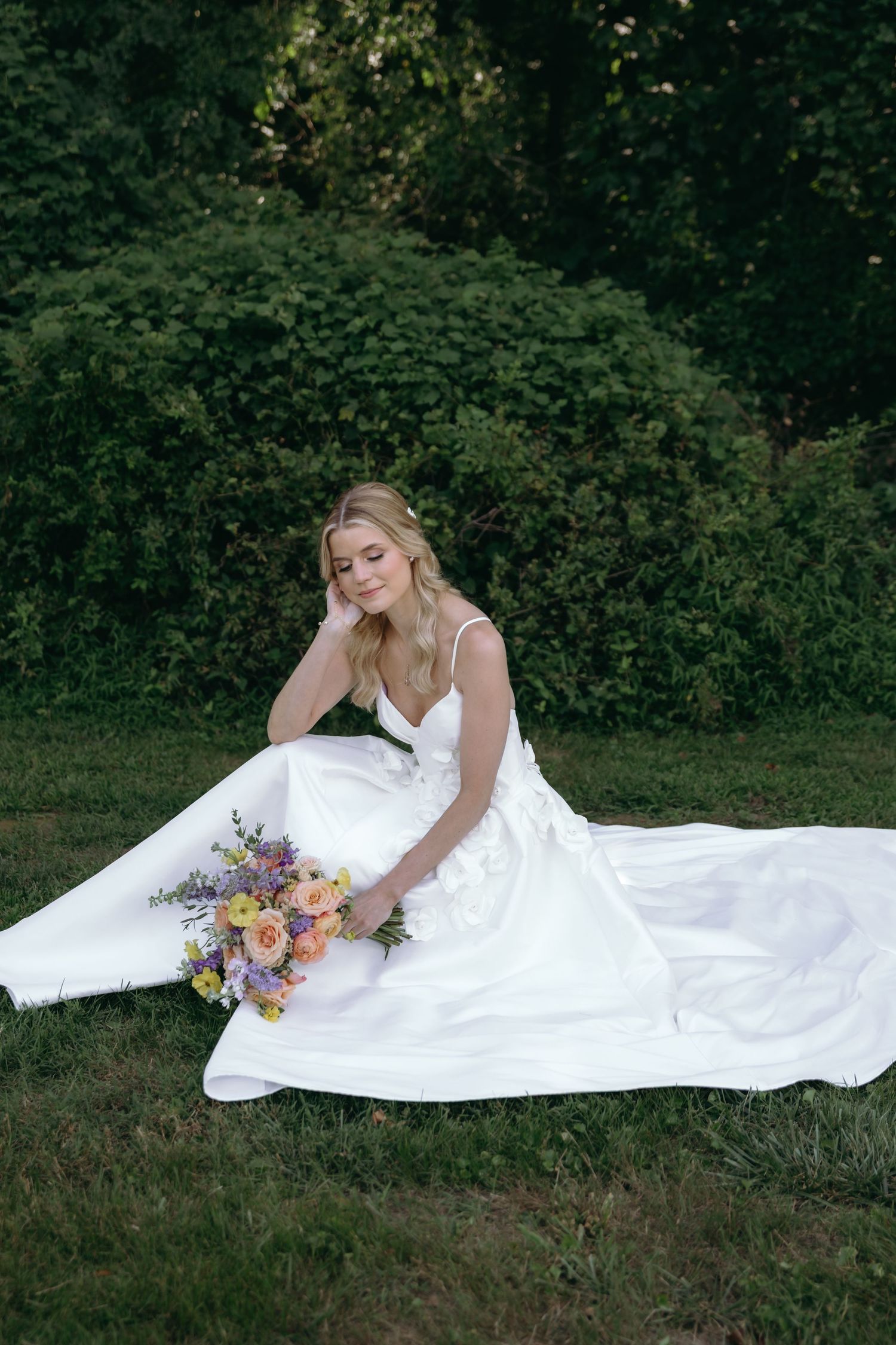 A bride in a flowing white gown poses gracefully on grass with a colorful floral bouquet.