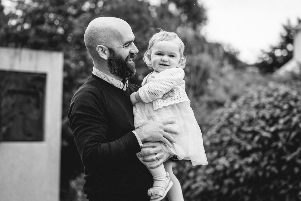 Black and white portrait of parent holding baby outdoors with greenery in background.