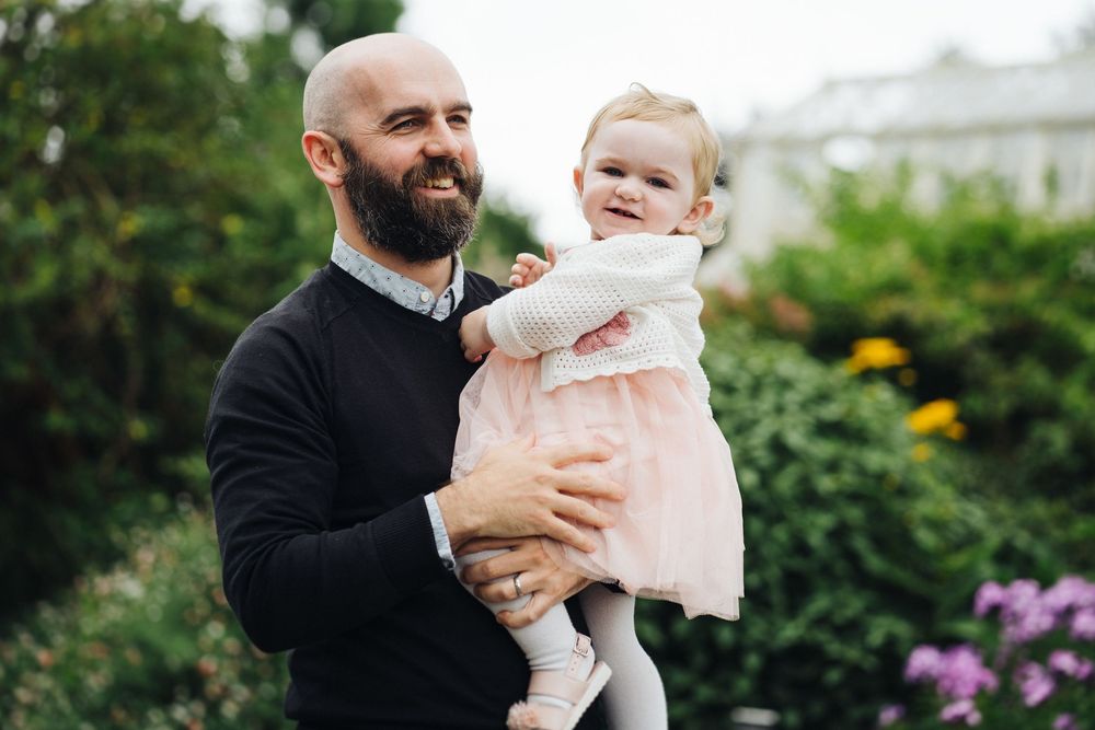 A parent wearing black holds a baby in a pink outfit while standing in a lush garden setting.