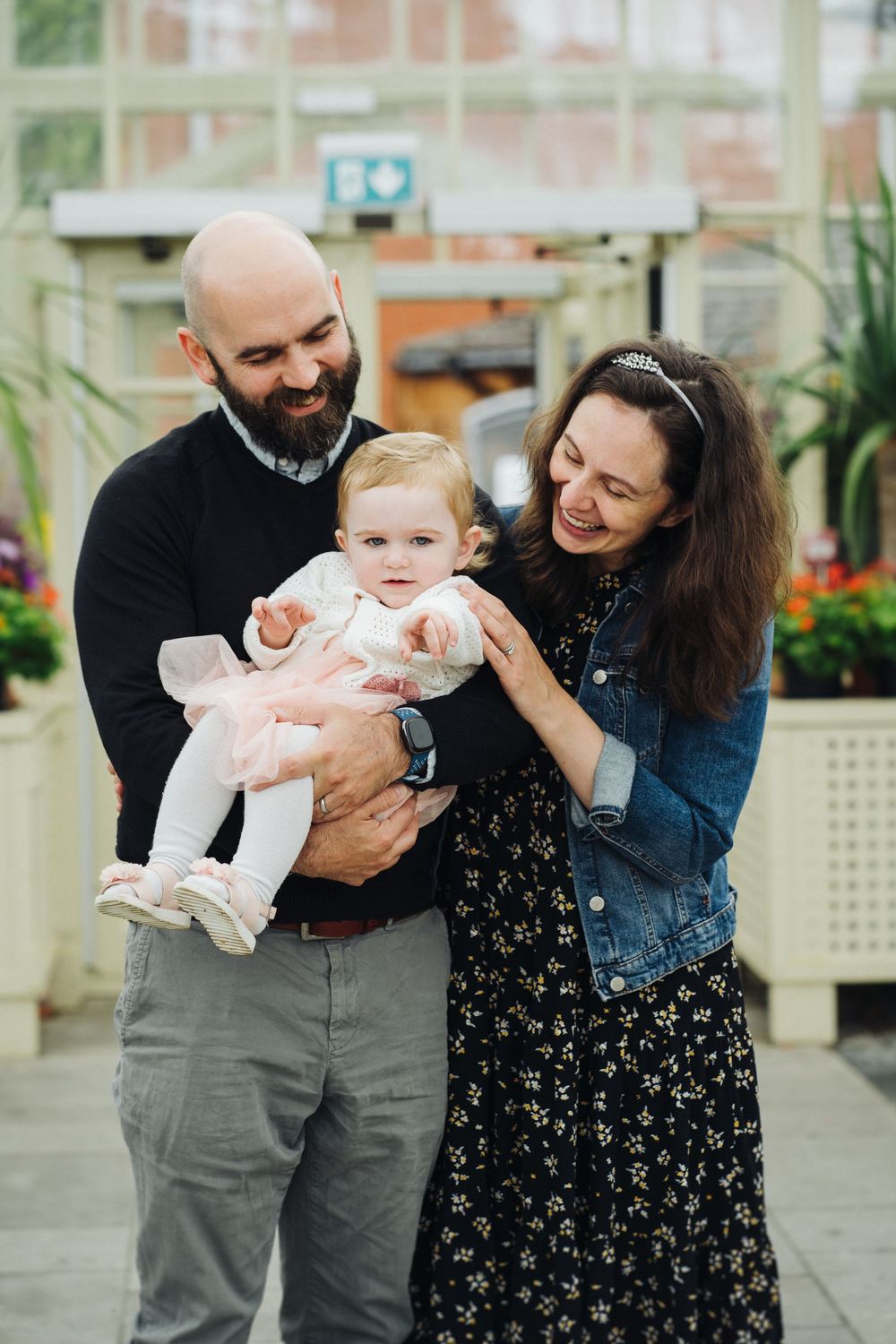 A couple in casual attire shares joyful moments with a baby in a greenhouse setting.