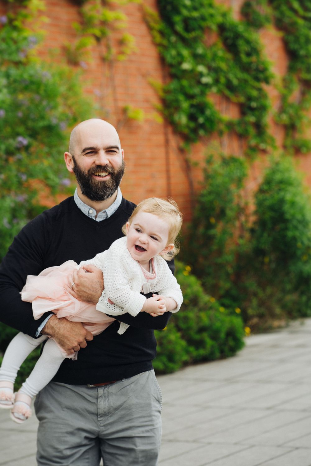 A person in a black sweater plays with a baby wearing white against a brick wall with green foliage background.