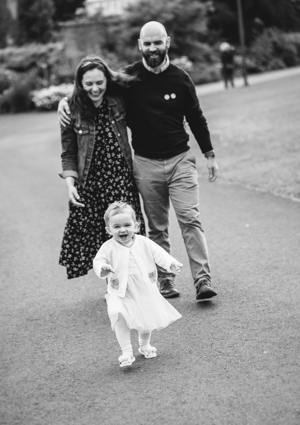 A family plays together in a park as a toddler in white takes first steps on a grassy slope.