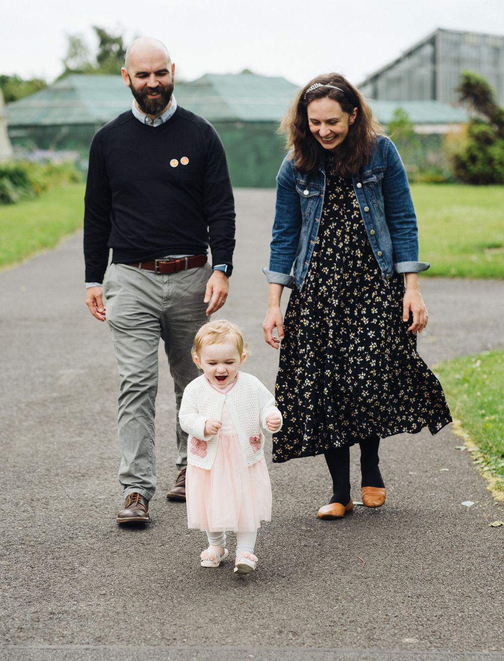 A family walks together on a paved path, with a small child in a pink dress taking steps between adults.