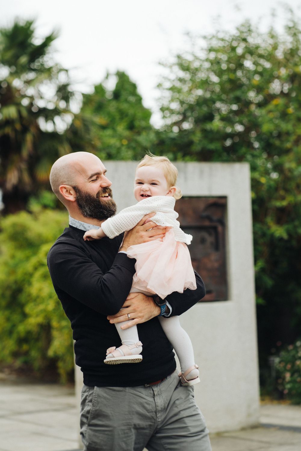 A bearded man in a black sweater joyfully holds a smiling baby in white clothing outdoors.