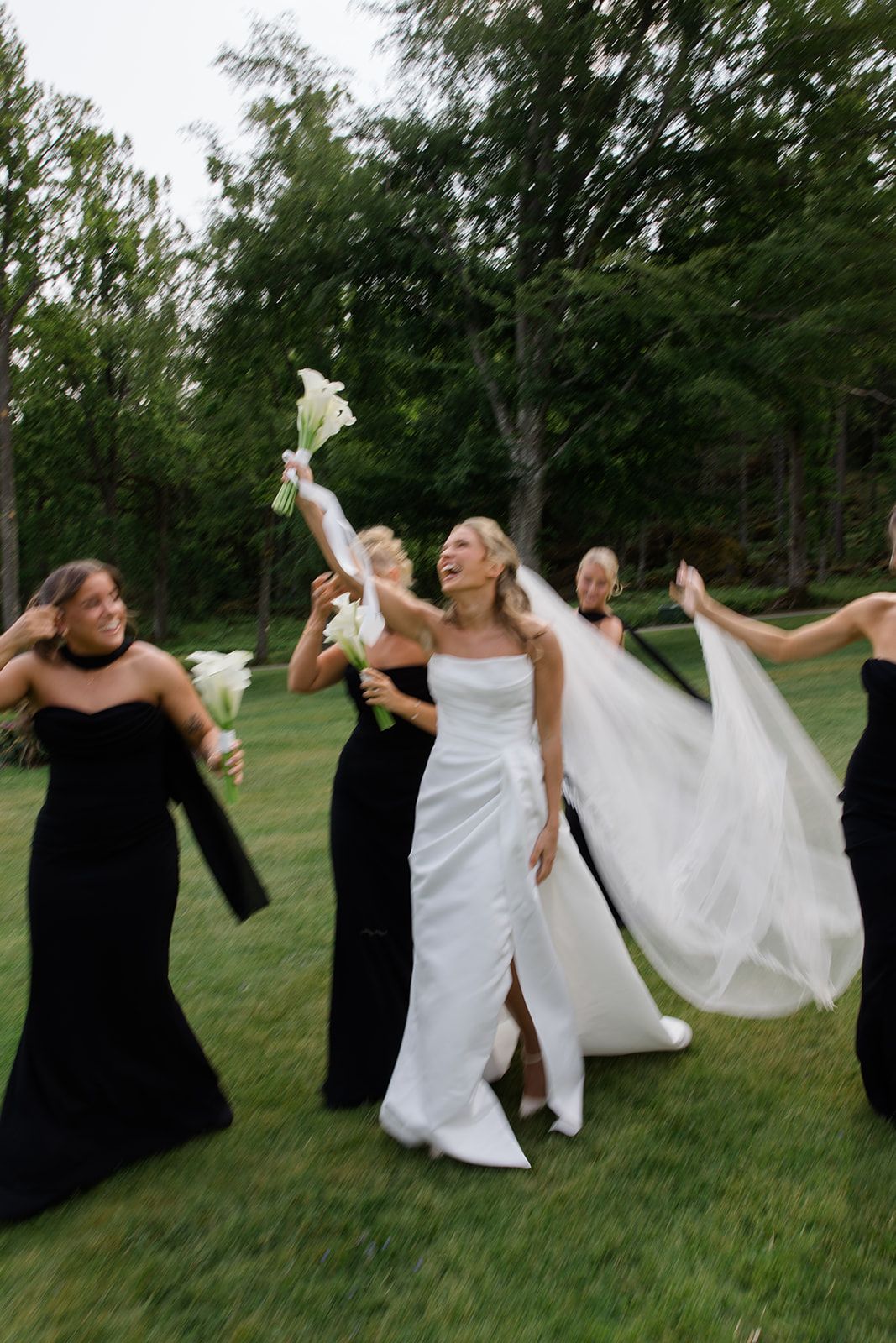 Bridesmaids in black dresses and bride in white gown pose playfully on grassy lawn in sequence of motion shots.