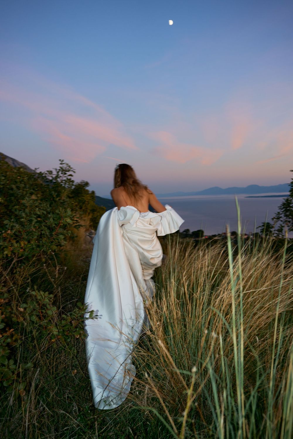 A bride in a flowing white dress stands against a coastal landscape at dusk with the moon visible.