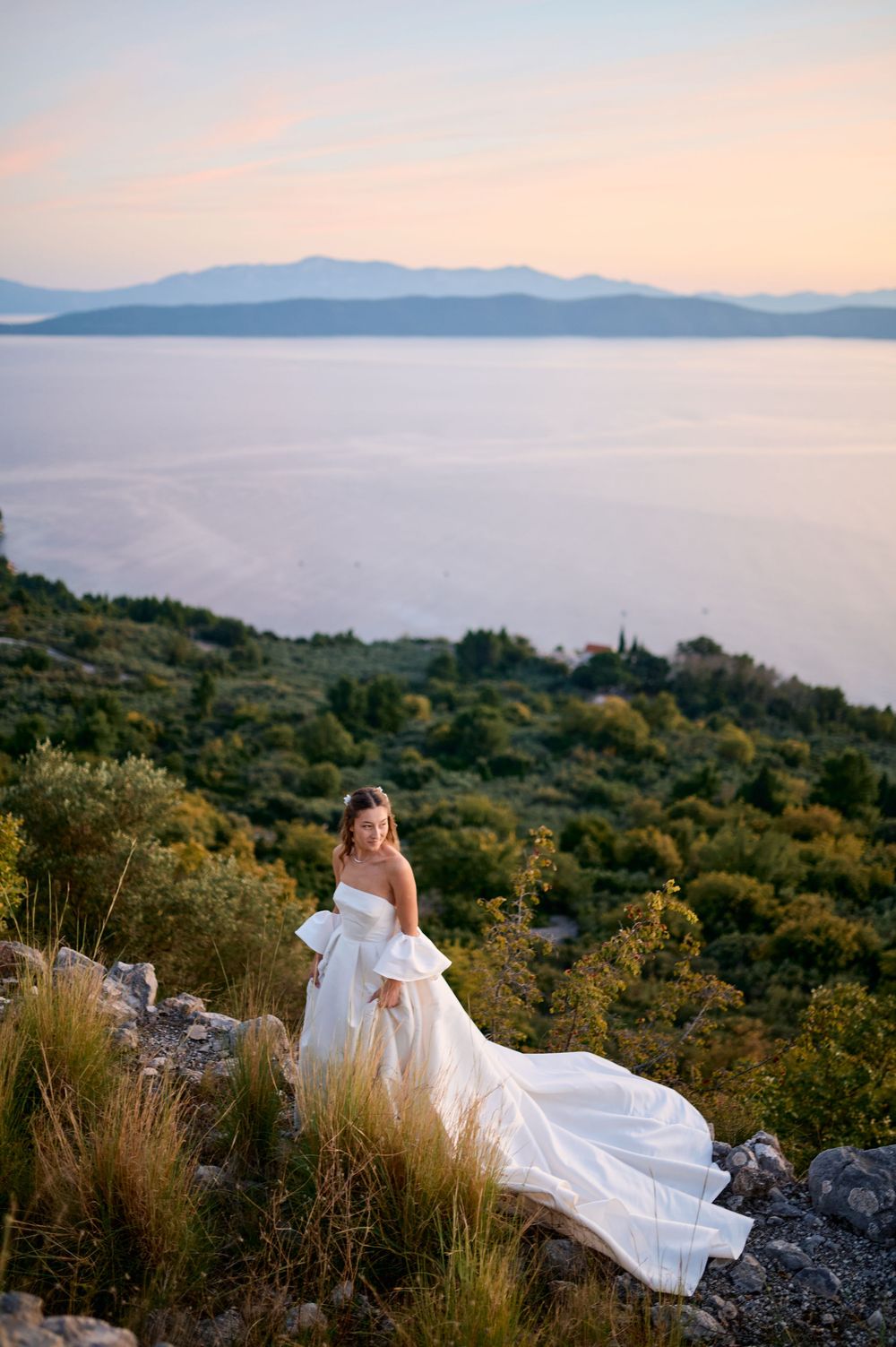 A bride in white rests on rocky terrain overlooking a scenic coastal vista at sunset.