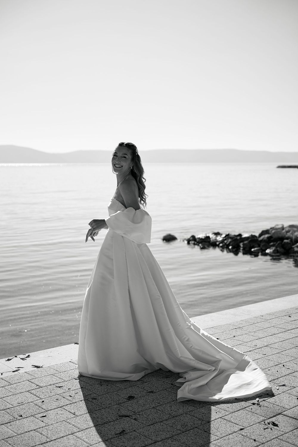 Black and white photograph of a bride in an elegant dress sitting by the seaside.
