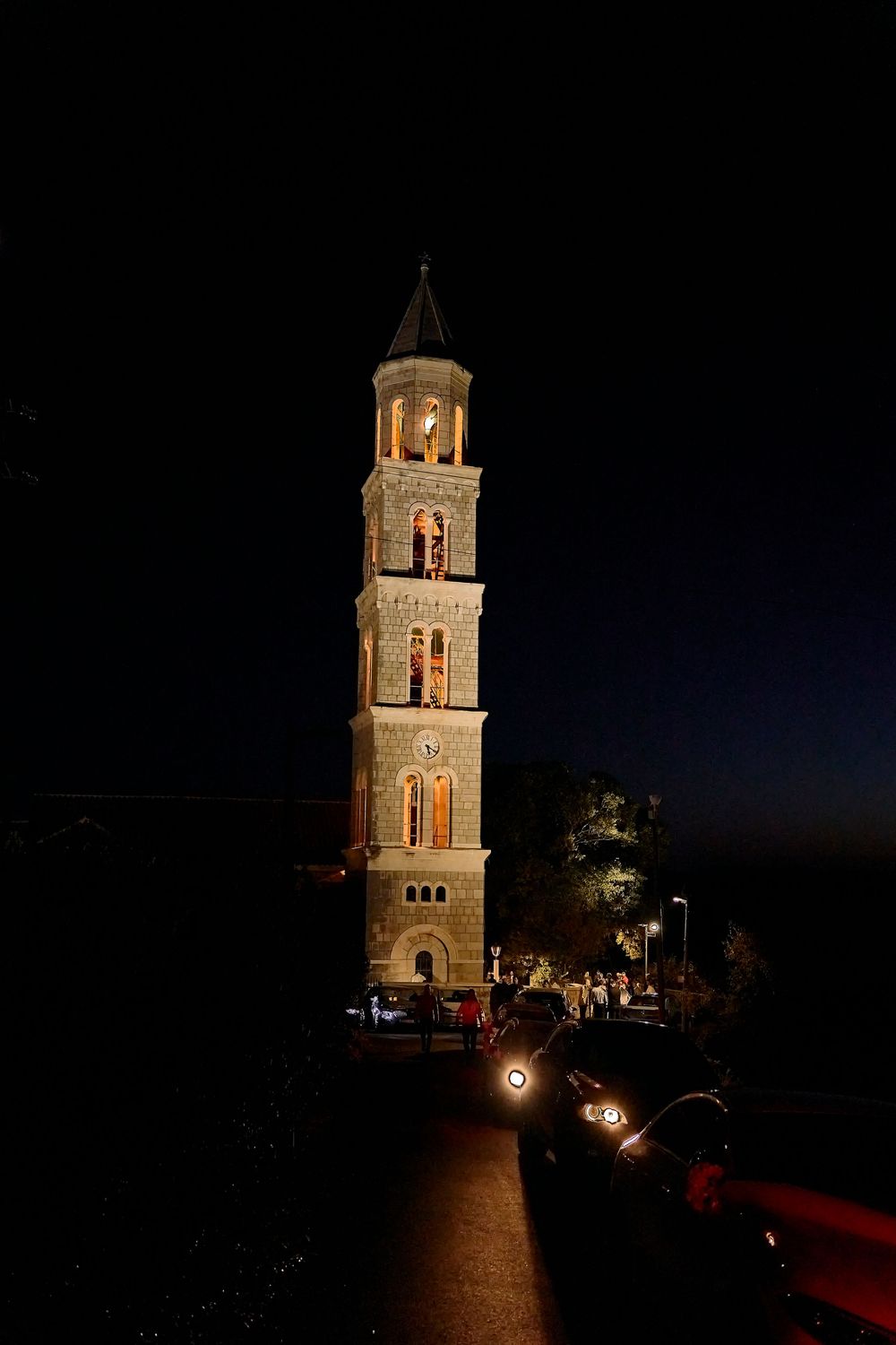 Illuminated church bell tower photographed at night against a dark sky with cars parked below.