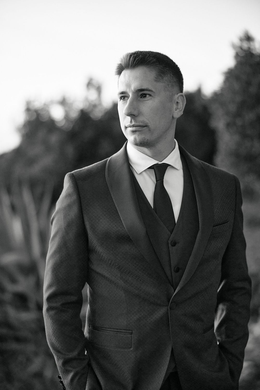 Black and white portrait series of groom in classic dark suit and tie against natural background.