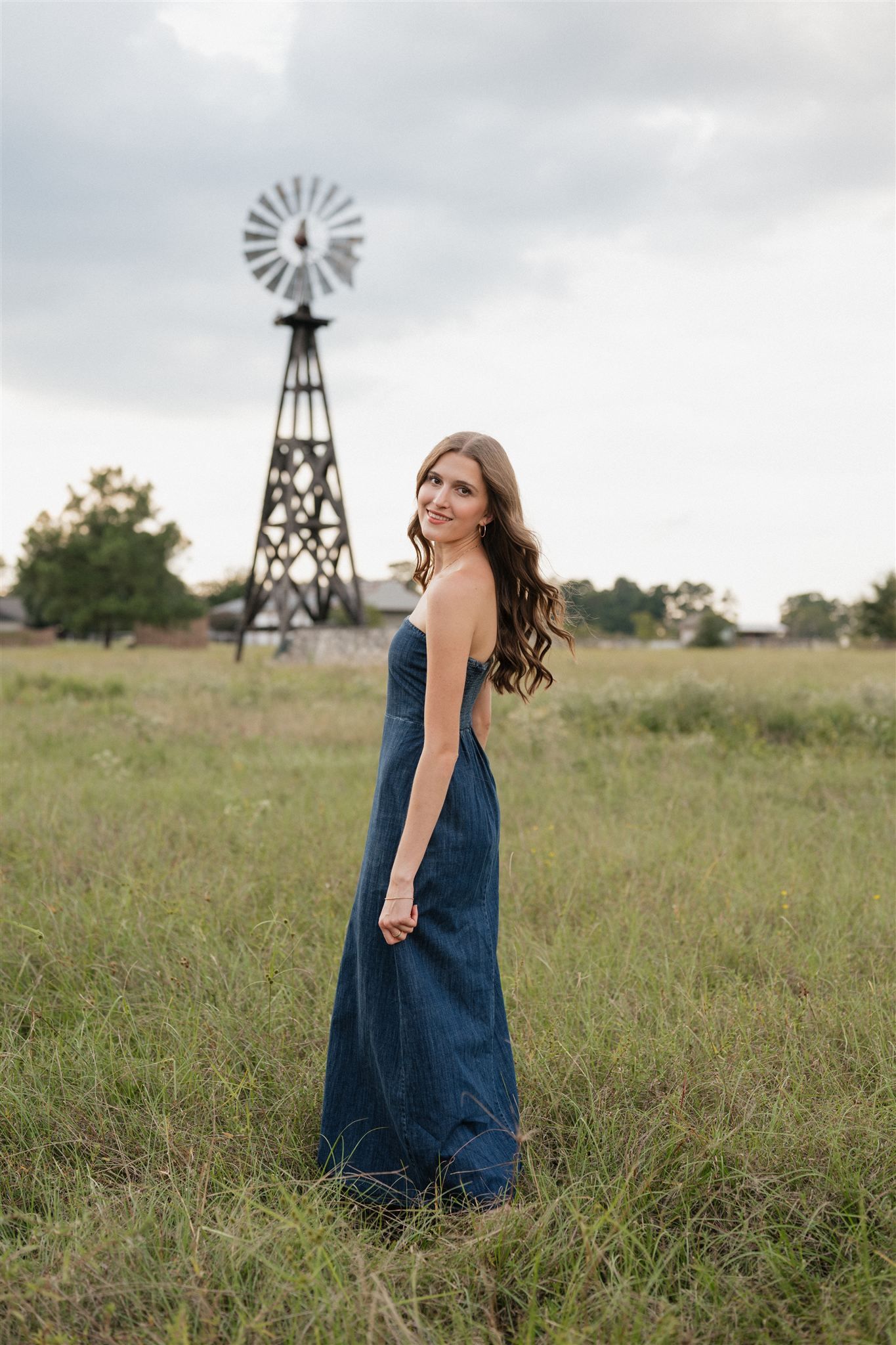 The Woodlands High School senior girl in a denim dress posing for pictures in Magnolia, Tx with a vintage windmill tower in the background.