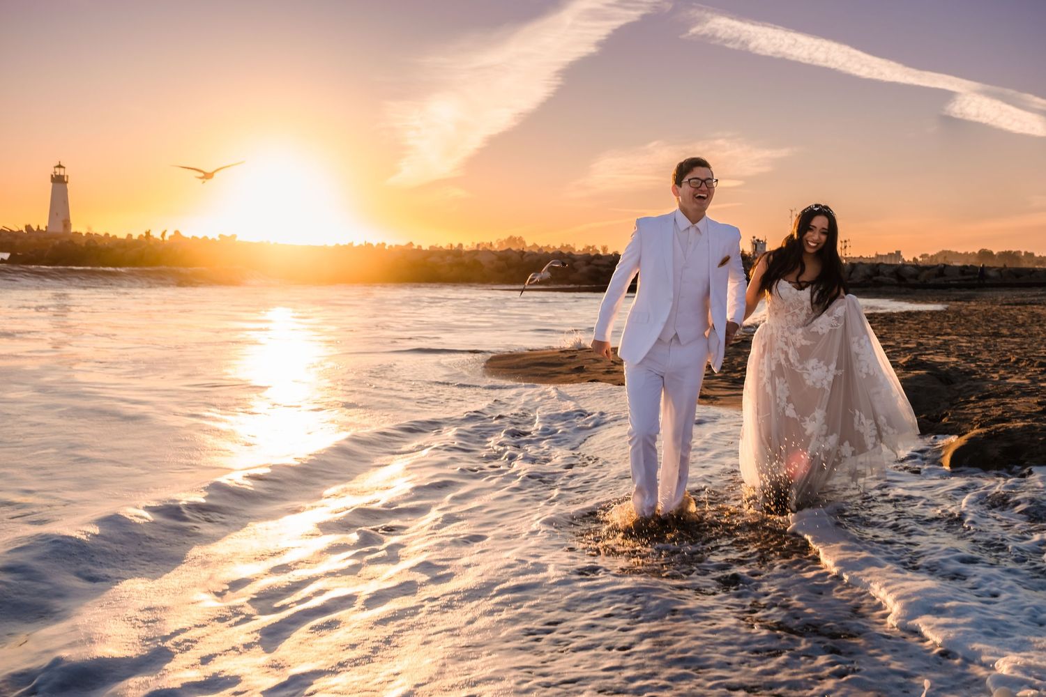 Two people walk along a rocky beach at sunset with a lighthouse silhouetted in the golden sky. Santa Cruz california wedding photos.