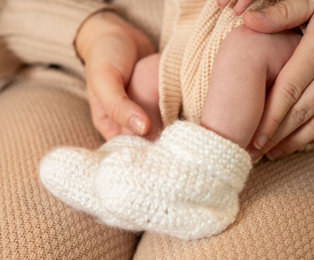 Hands placing a white crocheted baby bootie on a beige carpet surface.
