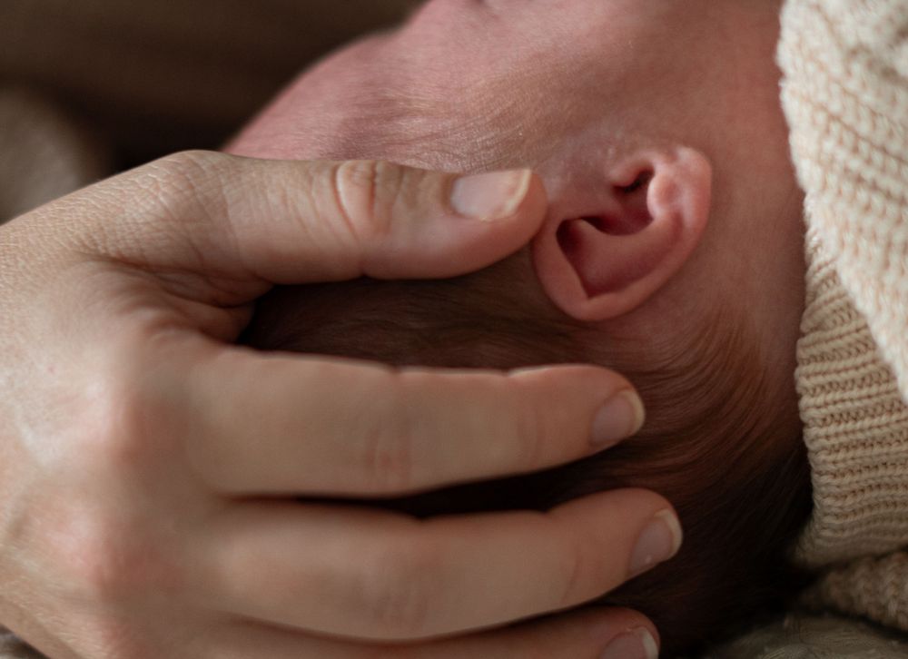 A gentle hand tenderly touches the ear of a newborn baby wearing a cream knit sweater.