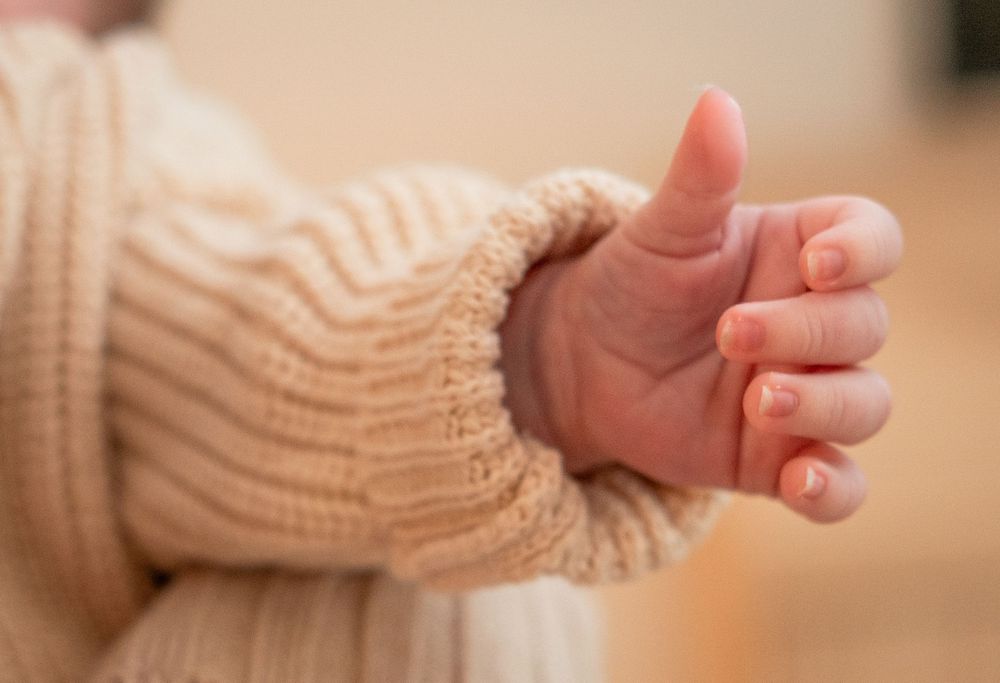 A baby wearing a knit sweater reaches out with their tiny hand against a warm beige background.