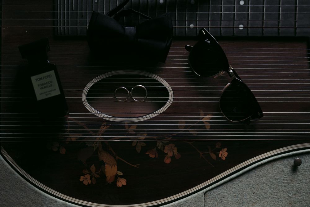 Two wedding rings sit on a dark acoustic guitar surface with sunglasses nearby in a moody artistic composition.