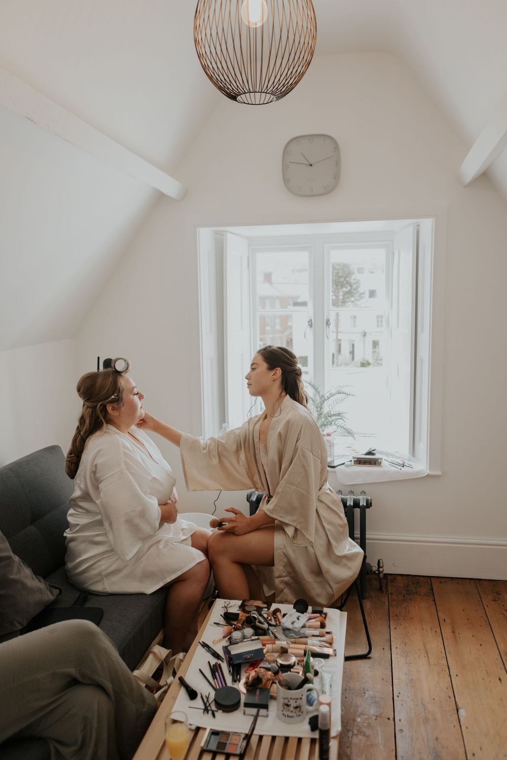 Makeup artist applying cosmetics to a bride seated by a bright window, with makeup spread on table.