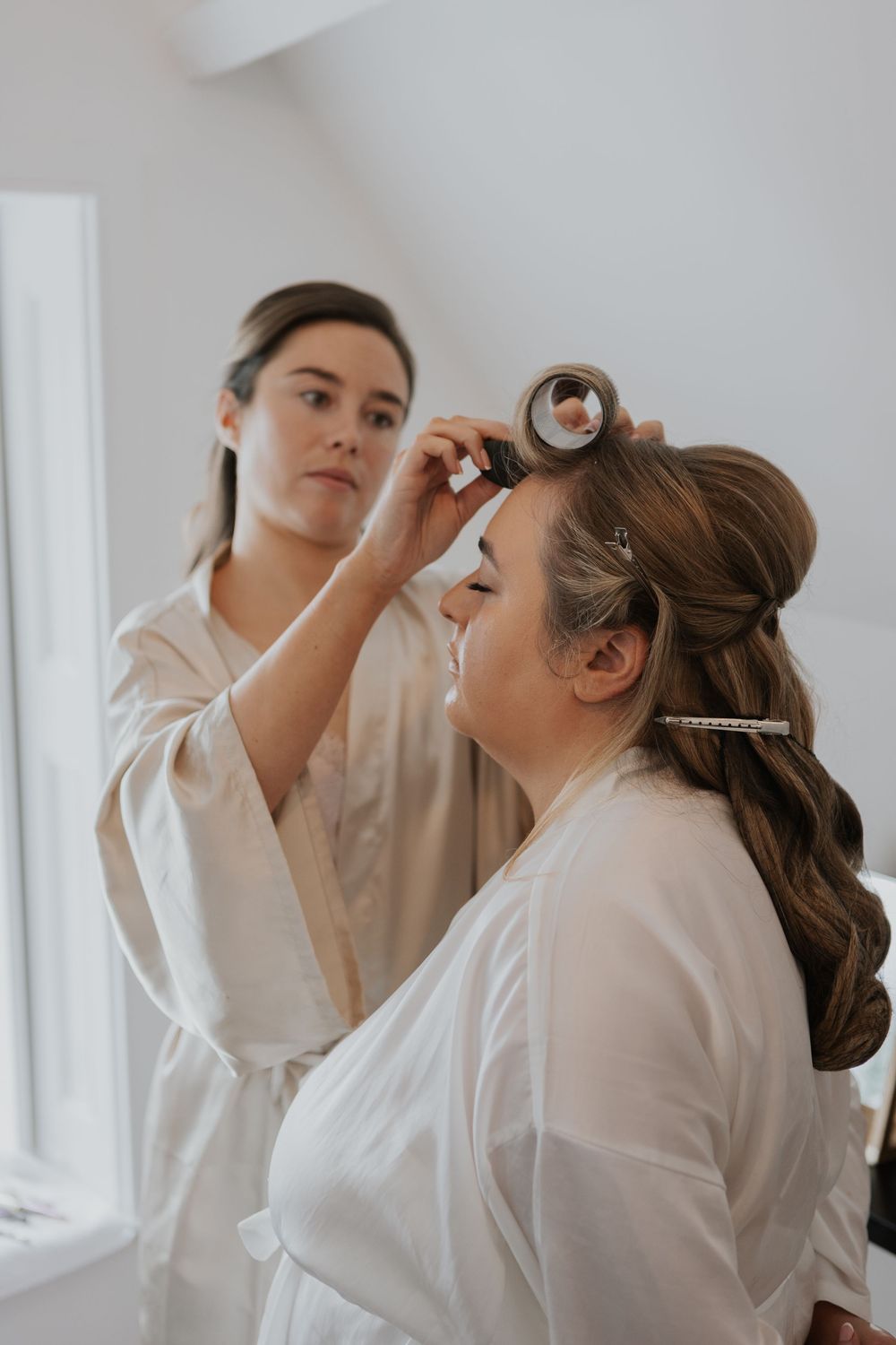 Hairstylist placing a roller in brunette bride's hair during wedding prep, both wearing white robes.