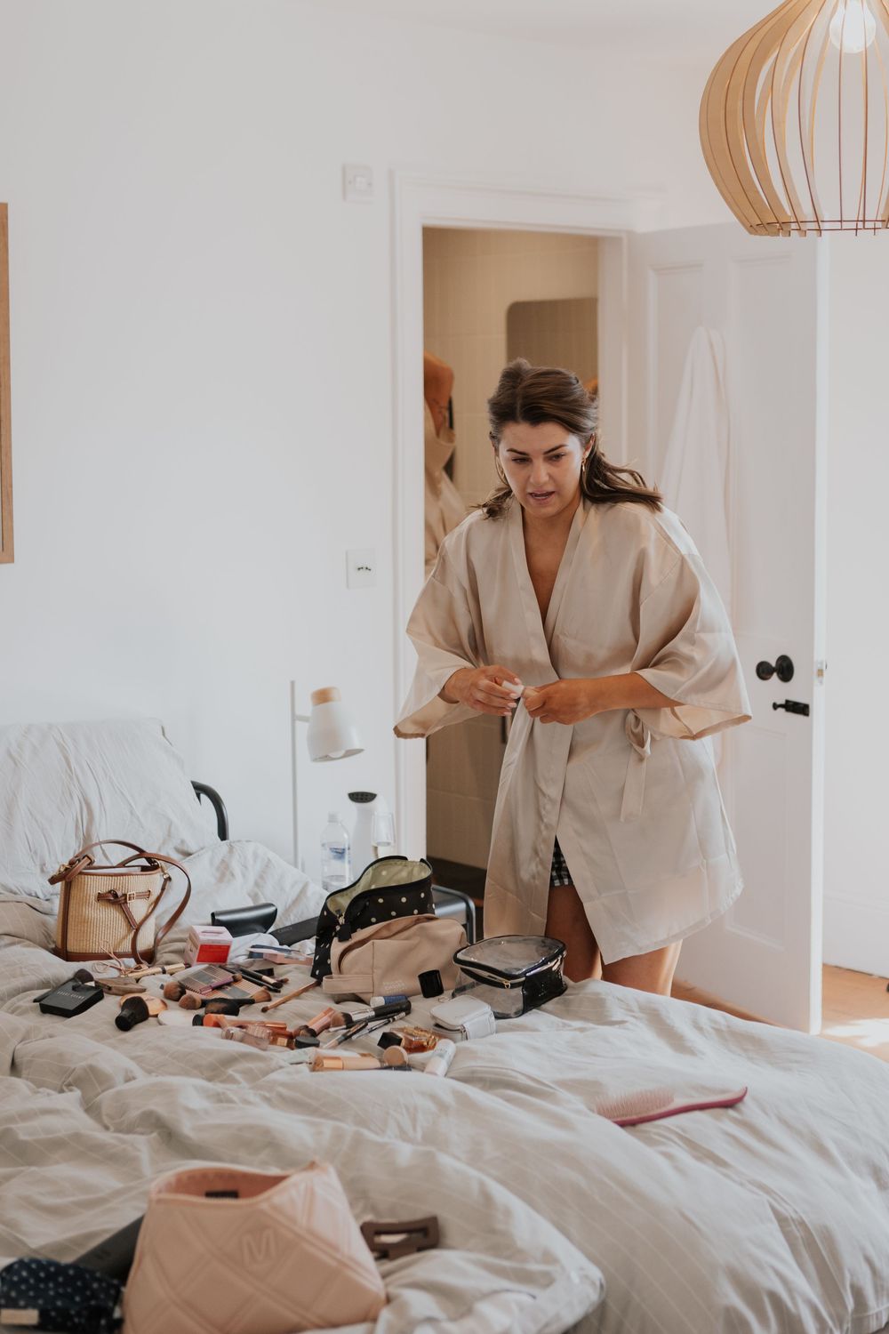 Bride in a beige robe organizing makeup and accessories spread across a white bed before her wedding.