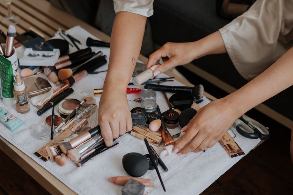 Close-up of hands selecting makeup products from a large collection of cosmetics on a table.