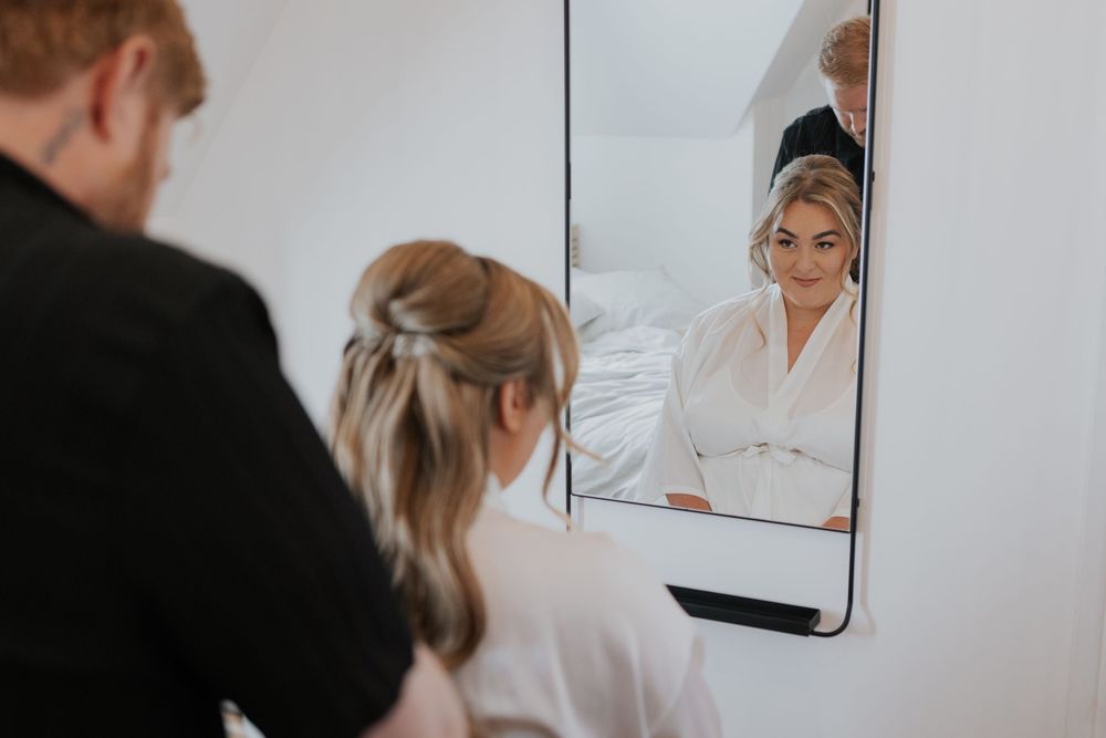 Bride smiling in mirror reflection while hairstylist works on her updo during wedding preparations.