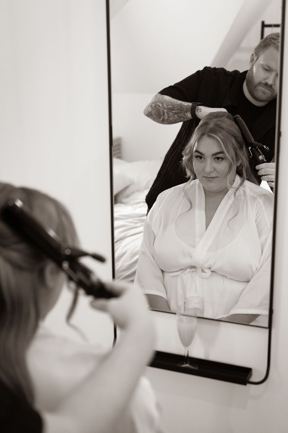 Black and white photo of tattooed hairstylist styling blonde bride's hair reflected in a mirror.