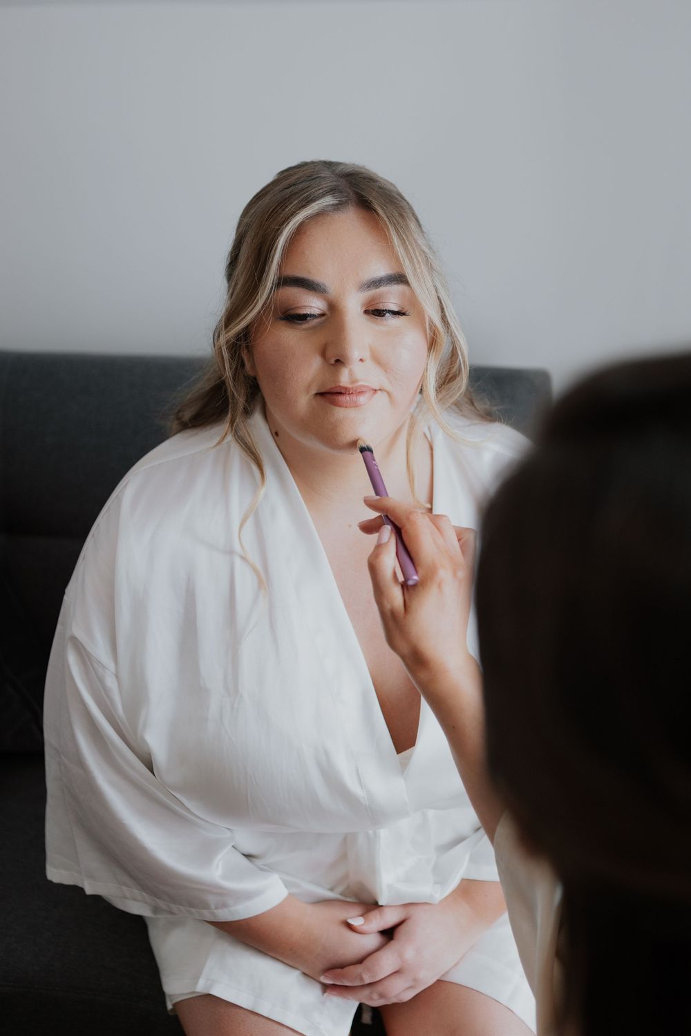 Makeup artist applying lip color to a blonde bride in a white robe during bridal preparations.
