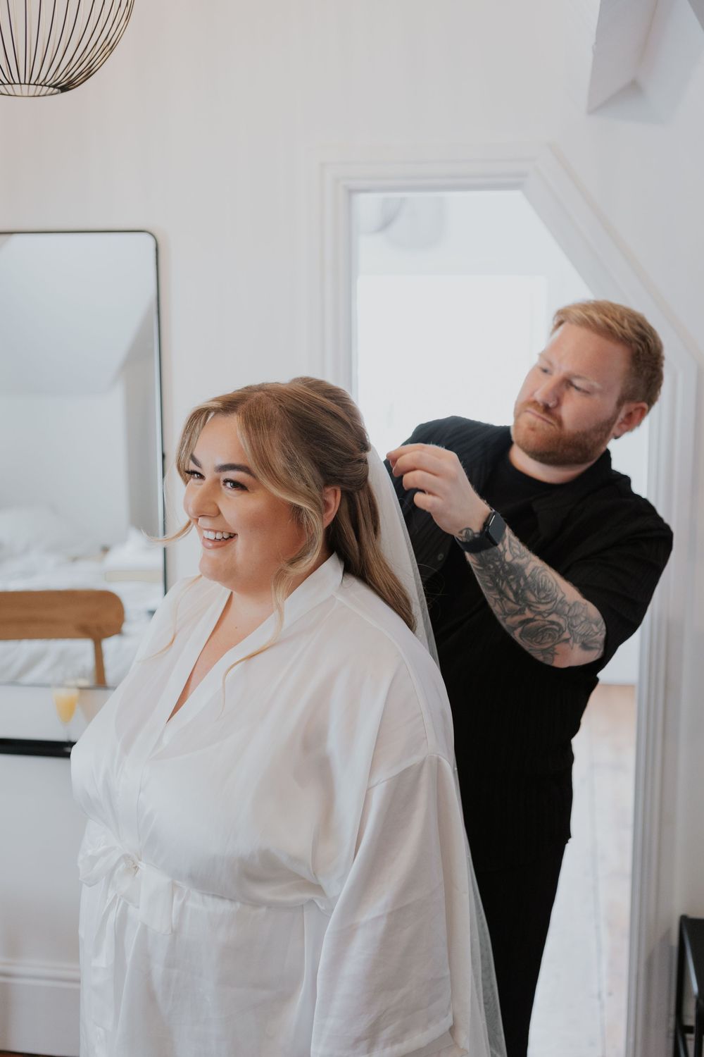 Tattooed male hairstylist styling smiling blonde bride's hair during wedding morning preparations.