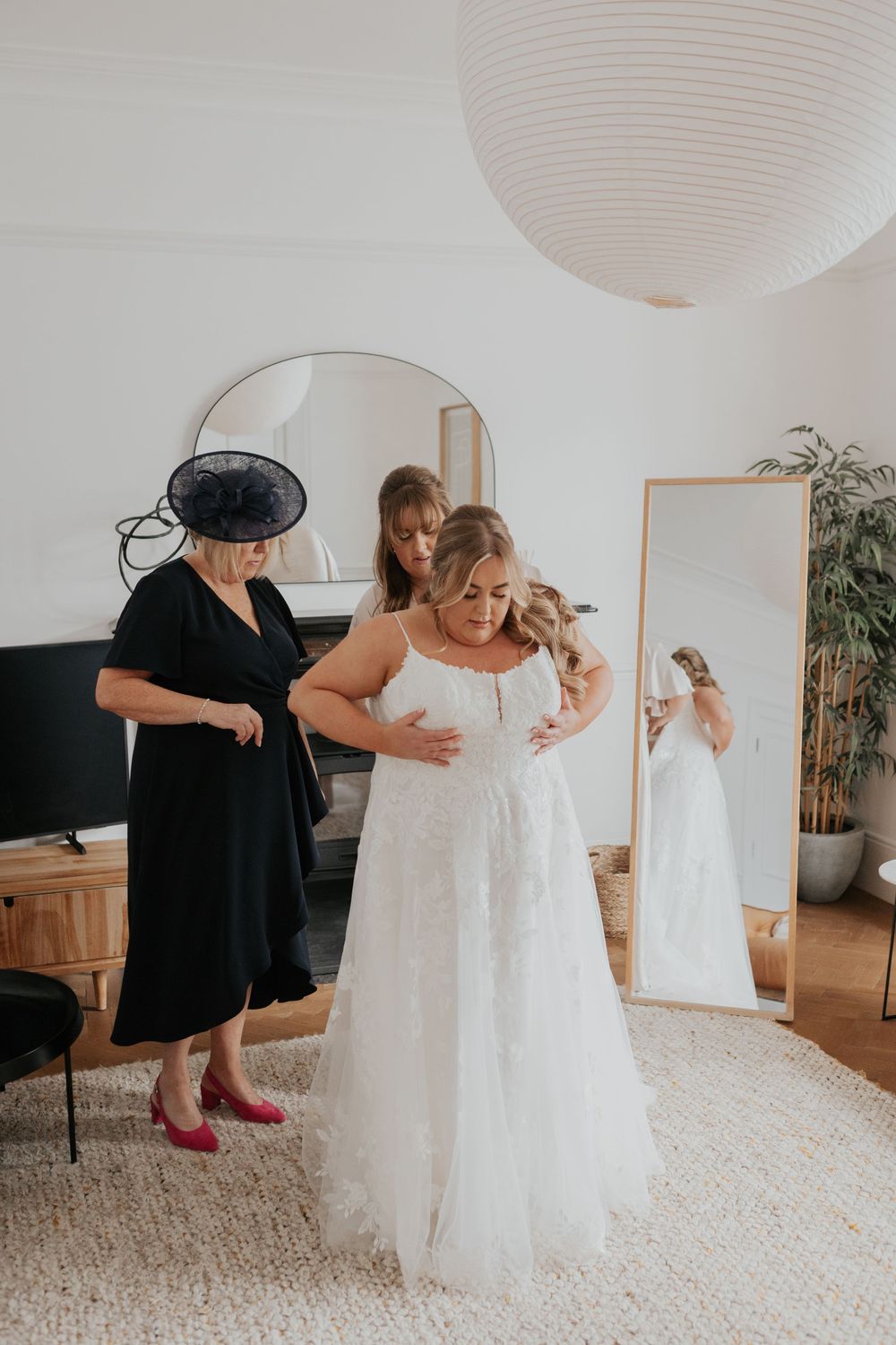 Bride smiling as two women help her into a white lace wedding dress in a bright modern room.
