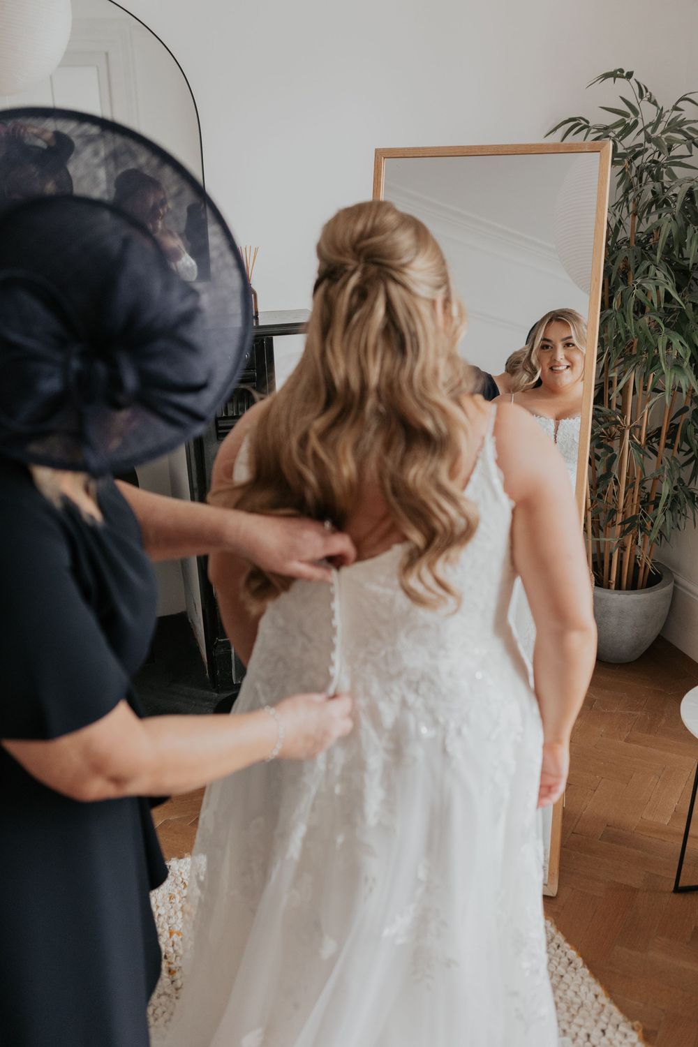 Woman in black fascinator buttoning the back of a bride's lace wedding gown near a mirror.