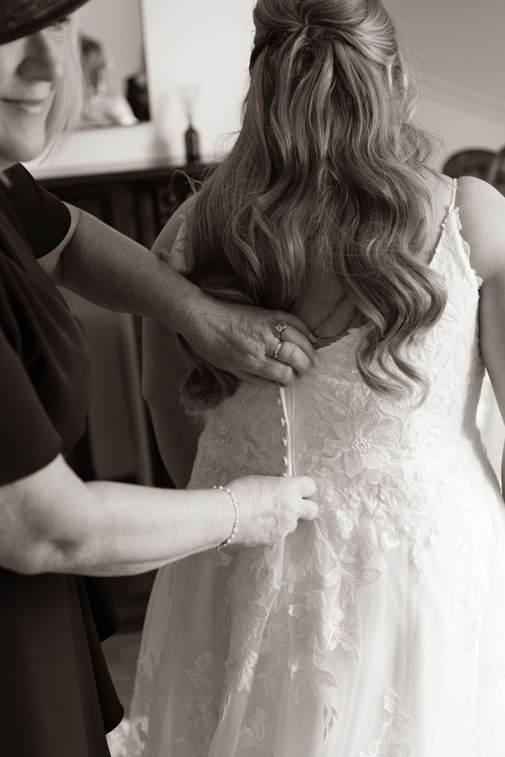 Black and white photo of hands fastening buttons on the back of a bridal lace wedding gown.