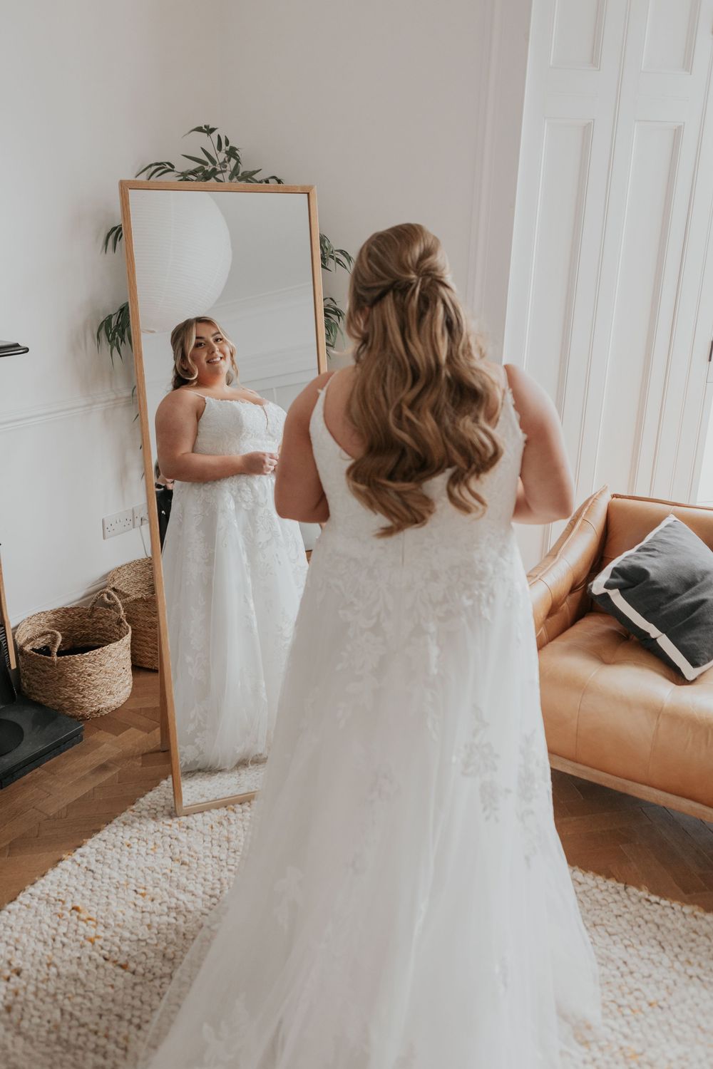 Smiling bride in a lace wedding dress admiring her reflection in a full-length wooden mirror.