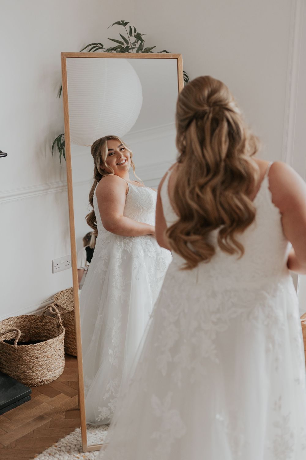 Bride in a floral lace wedding gown smiling joyfully at her reflection in a tall mirror.