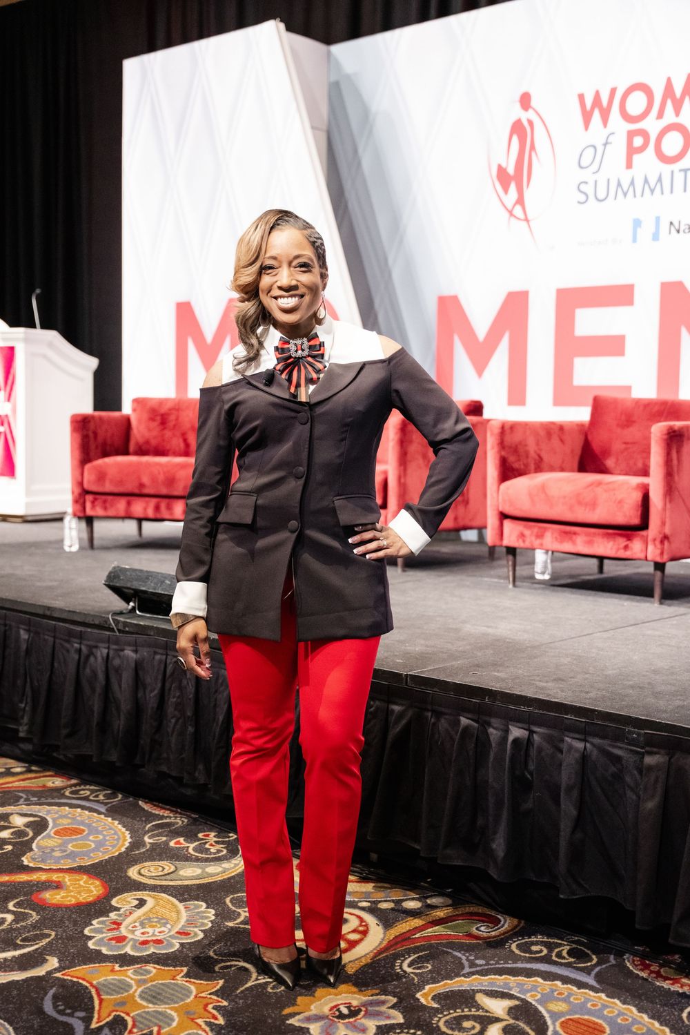 A person in a black jacket and red pants stands on a patterned carpet at a professional event with red signage.