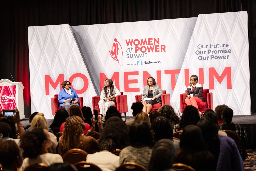 A large audience sits in a conference hall with red and white banners displaying 'Women' and 'Power' text on stage.