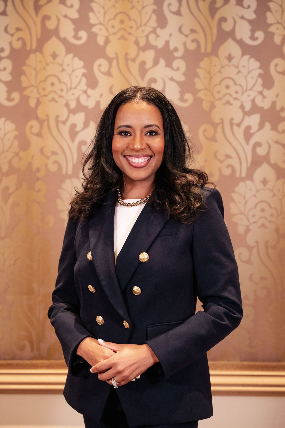 Professional wearing a navy blazer with gold buttons poses against an elegant damask wallpaper background.