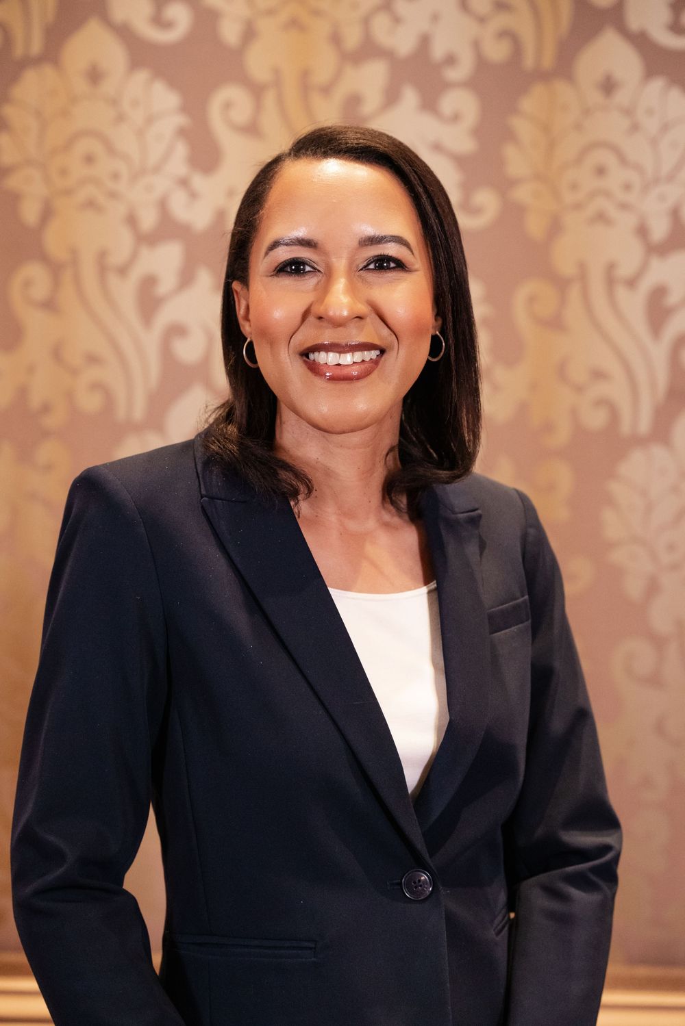 Professional wearing a black blazer and white top poses against a patterned gold wallpaper background.