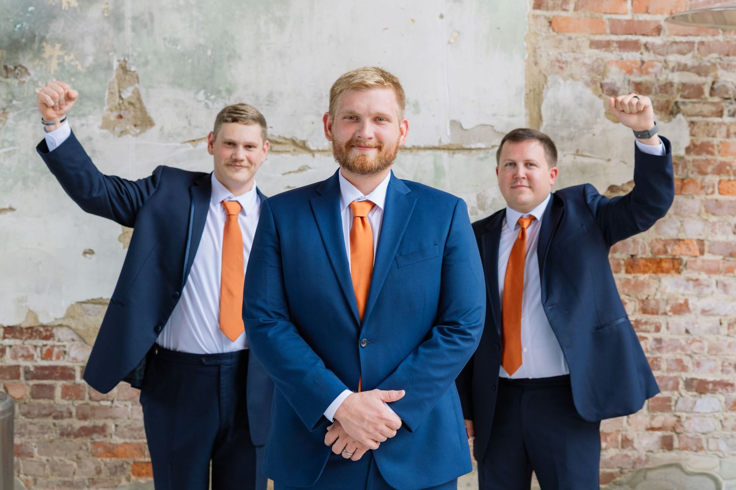 Groomsmen in navy suits and orange ties pose playfully against rustic brick wall at wedding venue.